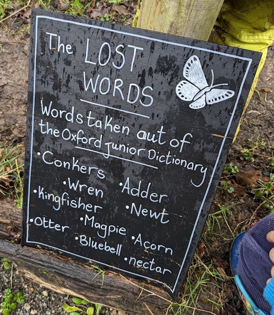 A sign outside in front of some mud and grass saying "The Lost Words: Words taken out of the Oxford Junior Dictionary. Conkers. Adder. Wren. Newt. Kingfisher. Magpie. Acorn. Otter. Bluebell. Nectar.